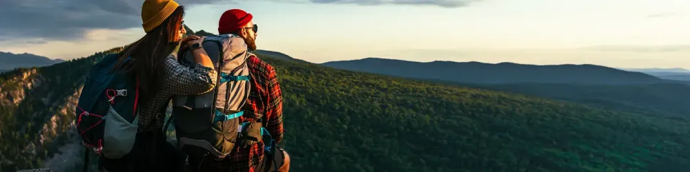 The couple meets the sunset in the mountains. Two travelers are sitting on the edge of a cliff and admiring the beautiful panoramic view. Travelers with backpacks at sunset in the mountains. Panorama